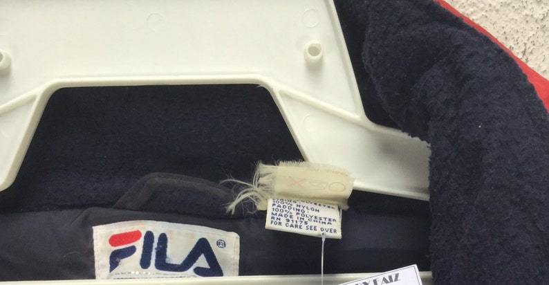 May include: Close-up of a navy blue and red FILA jacket. The jacket is hanging on a white hanger. The FILA logo is visible on the jacket. A tag with care instructions is also visible.