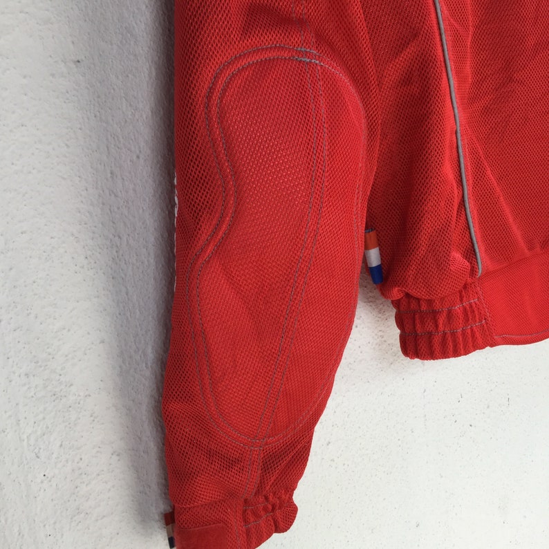 May include: Red mesh jacket with a zipper closure and a white trim. The jacket has a patch on the left sleeve.