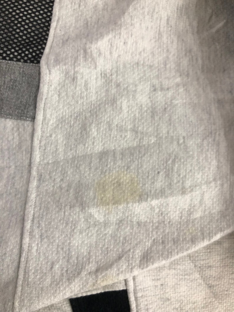 May include: Close-up of a light gray fabric with a visible stain. The fabric appears to be a soft material, possibly a sweatshirt or similar garment. The image also shows a section of black mesh and gray fabric.