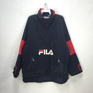 May include: A navy blue Fila anorak jacket with red accents on the sleeves and collar. The jacket features a white Fila logo on the front, a button-up closure, and two zippered pockets. The jacket is hanging on a white hanger.