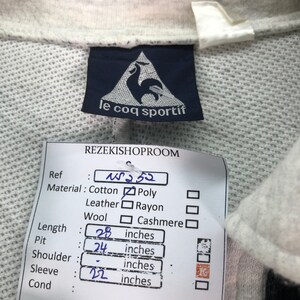 May include: A close-up of a light gray Le Coq Sportif sweatshirt tag. The tag is navy blue with a white rooster logo. A price tag indicates the length is 28 inches, pit is 24 inches, and sleeve is 22 inches.