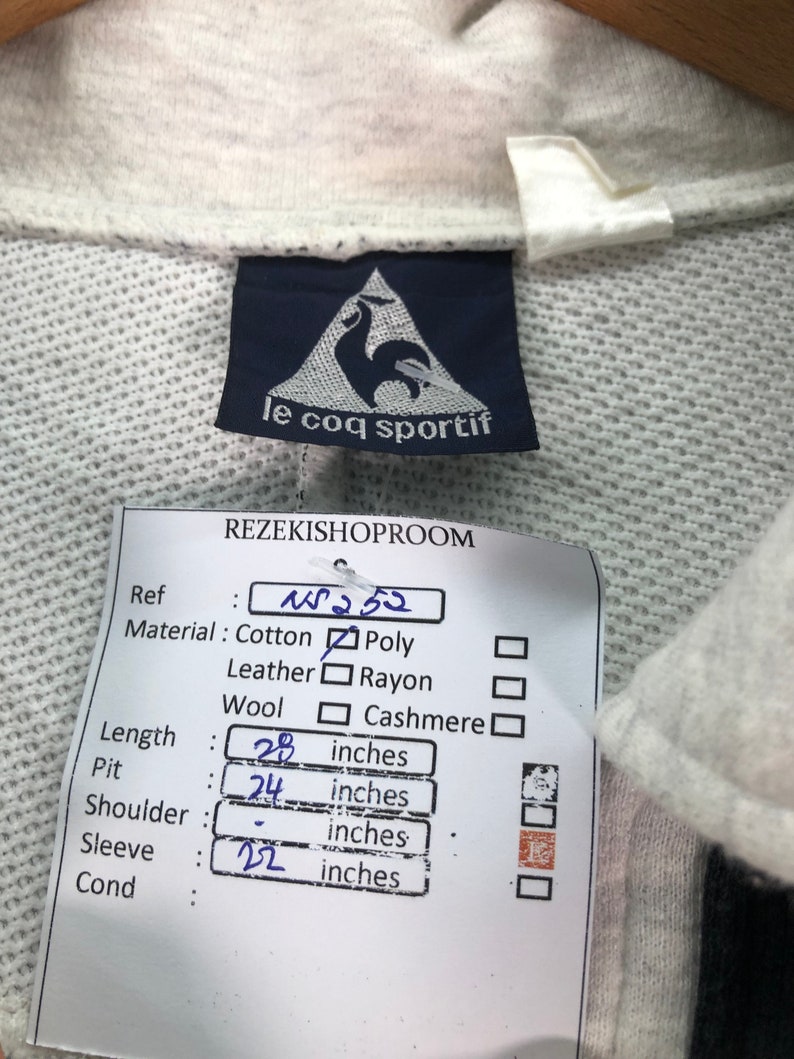 May include: A close-up of a light gray Le Coq Sportif sweatshirt tag. The tag is navy blue with a white rooster logo. A price tag indicates the length is 28 inches, pit is 24 inches, and sleeve is 22 inches.