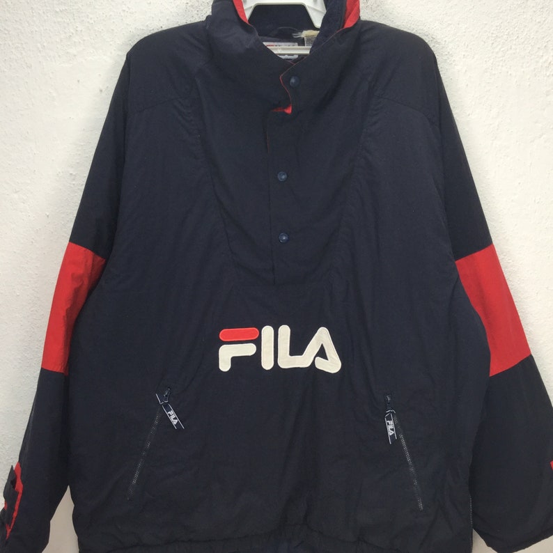 May include: A navy blue Fila anorak jacket with red accents on the sleeves. The jacket features a high collar, a button-up front, and two zippered pockets. The Fila logo is prominently displayed in white across the chest.