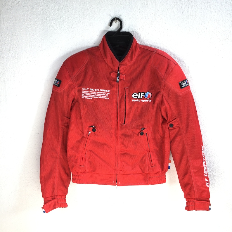 May include: Red motorcycle jacket with a zipper closure and black trim. The jacket has the text "ELF MOTO RANGE" and "ELF moto sports" printed on the front. The jacket also has the text "ELF COMPETITION" printed on the right sleeve.