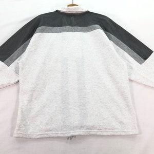 May include: A long-sleeved sweatshirt in a light gray color, featuring a color block design across the shoulders and sleeves. The color block includes shades of gray. A small red logo is visible on the sleeve.