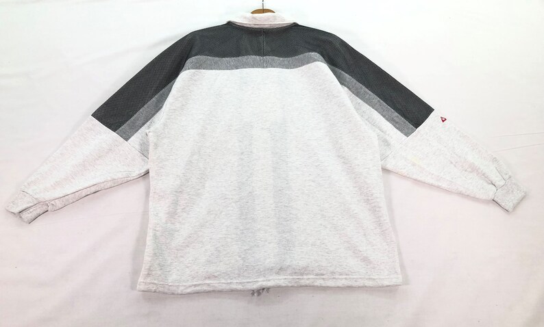 May include: A long-sleeved sweatshirt in a light gray color, featuring a color block design across the shoulders and sleeves. The color block includes shades of gray. A small red logo is visible on the sleeve.