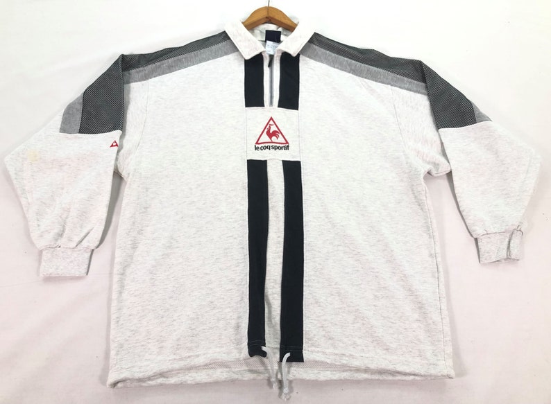 May include: A light gray long-sleeved sweatshirt with a collar and a quarter-zip closure. The shirt features black and gray accents on the shoulders and a black stripe down the front. The Le Coq Sportif logo is on the chest.