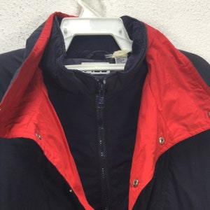 May include: A navy blue jacket with a red collar and accents, hanging on a white hanger. The jacket has a full zipper and snap closures. The collar is lined with a soft, dark blue material.