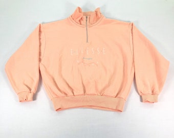 May include: A peach-colored sweatshirt with a high collar and a quarter-zip closure. The brand name "ellesse" is embroidered in white, with "soruggio" below. The cuffs and hem are ribbed.