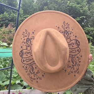 May include: A tan wide-brimmed hat featuring a dark brown printed design of two snakes intertwined with leafy branches and small starburst motifs. The hat has a structured crown with a central indentation and a flat, circular brim.