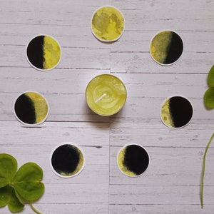 May include: A set of eight yellow and black moon phase stickers on a white wooden surface. The stickers are arranged in a circle around a small white candle.