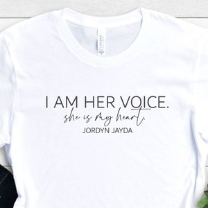 May include: White t-shirt with the text "I AM HER VOICE. she is my heart" and the name "JORDYN JAYDA" printed in black. The shirt is folded, with a pair of dark blue jeans visible below. A small green plant is in the background.