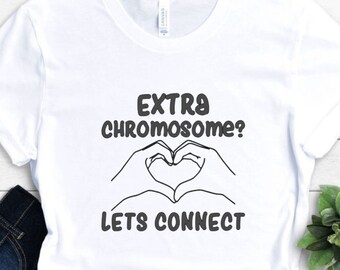 Könnte beinhalten: Weißes T-Shirt mit dem schwarzen Text "EXTRA CHROMOSOME? LETS CONNECT". Das Design zeigt ein Herz, das von zwei Händen gebildet wird. Das T-Shirt ist mit einer blauen Jeans und einer kleinen grünen Pflanze gefaltet.