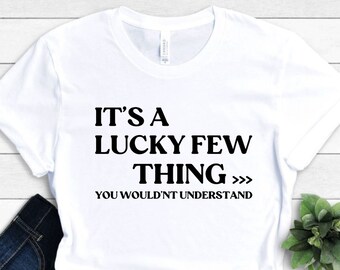May include: White t-shirt with the text "IT'S A LUCKY FEW THING YOU WOULDN'T UNDERSTAND" in black. The shirt is folded, with dark blue jeans visible. A small green plant is in the background.