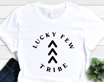 May include: White t-shirt featuring the text "LUCKY FEW TRIBE" in a black circular design. The design includes three upward-pointing arrows. The shirt is paired with dark blue jeans.
