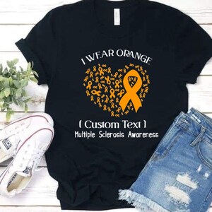 Op de afbeelding: Zwart T-shirt met de tekst "I Wear Orange" en een hart van oranje linten. Het shirt bevat de tekst "(Custom Text) Multiple Sclerosis Awareness". Witte sneakers en een spijkerbroekshort liggen naast het shirt.