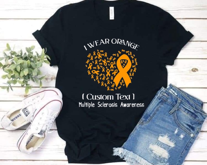 Op de afbeelding: Zwart T-shirt met de tekst "I Wear Orange" en een hart van oranje linten. Het shirt bevat de tekst "(Custom Text) Multiple Sclerosis Awareness". Witte sneakers en een spijkerbroekshort liggen naast het shirt.