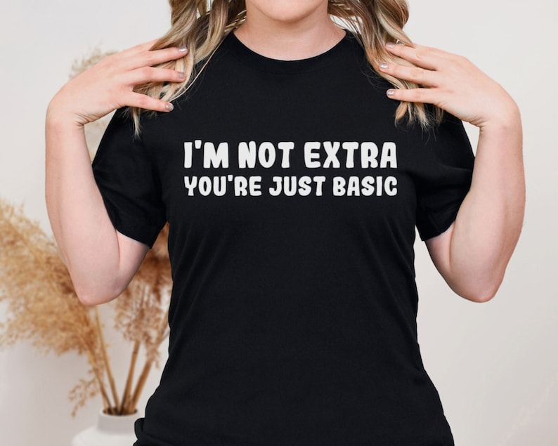 Pu&ograve; includere: T-shirt nero con la scritta bianca "I'M NOT EXTRA YOU'RE JUST BASIC". La maglietta &egrave; a girocollo e viene indossata da una persona. La persona tiene la maglietta sulle spalle.