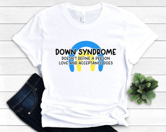 Puede incluir: Camiseta blanca con el texto "DOWN SYNDROME DOESN'T DEFINE A PERSON LOVE AND ACCEPTANCE DOES" en negro. El diseño incluye un arcoíris azul y amarillo. La camiseta está doblada, con unos vaqueros azul oscuro visibles.