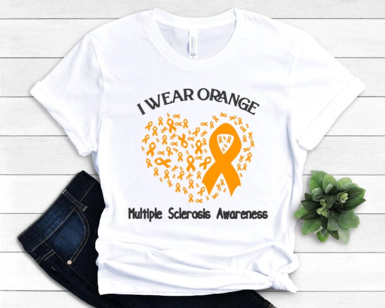 Op de afbeelding: Wit T-shirt met de tekst "I Wear Orange" boven een hartvormig ontwerp van oranje linten. De woorden "Multiple Sclerosis Awareness" staan eronder. Het shirt wordt getoond op een donkerblauwe jeans.