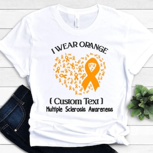 Op de afbeelding: Wit T-shirt met de tekst "I Wear Orange" boven een hartvormig ontwerp van oranje linten. De tekst "(Custom Text) Multiple Sclerosis Awareness" staat eronder. Het shirt wordt gepresenteerd op een witte ondergrond.