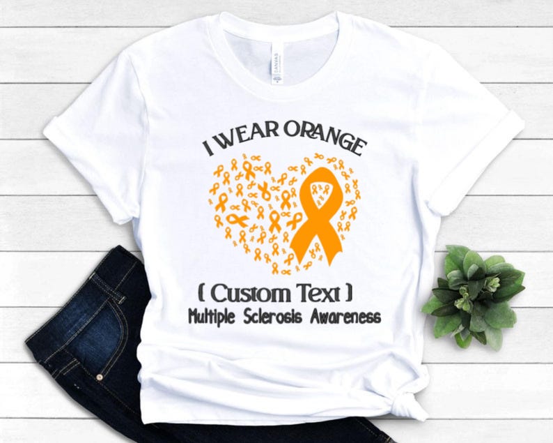 Op de afbeelding: Wit T-shirt met de tekst "I Wear Orange" boven een hartvormig ontwerp van oranje linten. De tekst "(Custom Text) Multiple Sclerosis Awareness" staat eronder. Het shirt wordt gepresenteerd op een witte ondergrond.