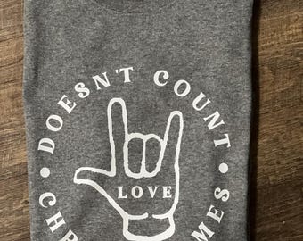 Könnte beinhalten: Ein gefaltetes graues T-Shirt mit einer weißen Grafik. Die Grafik zeigt eine Hand im Zeichen der Liebe, mit dem Wort "LOVE" in der Mitte. Die Worte "DOESN'T COUNT CHROMOSOMES" sind in einem Kreis um die Hand angeordnet.