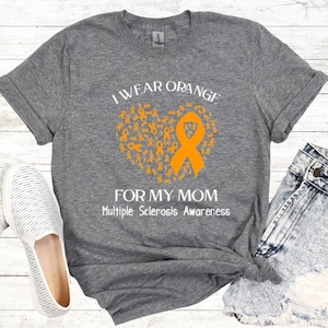 Op de afbeelding: Een grijs t-shirt met de tekst "I WEAR ORANGE FOR MY MOM" en "Multiple Sclerosis Awareness". Het ontwerp toont een oranje lint en een hartvorm gemaakt van oranje linten.