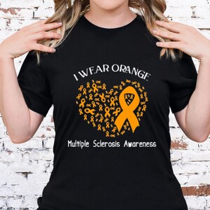 Op de afbeelding: Zwart T-shirt met de tekst "I Wear Orange" boven een hartvormig ontwerp van oranje linten. De woorden "Multiple Sclerosis Awareness" staan eronder.