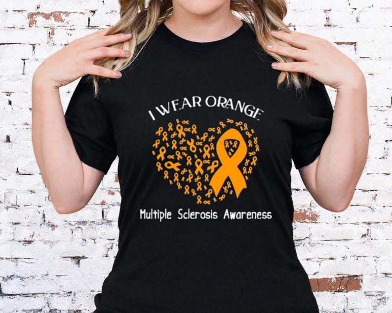Op de afbeelding: Zwart T-shirt met de tekst "I Wear Orange" boven een hartvormig ontwerp van oranje linten. De woorden "Multiple Sclerosis Awareness" staan eronder.