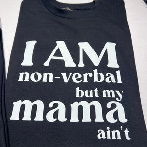 May include: Black t-shirt with white text that reads "I AM non-verbal but my mama ain't". The shirt is folded and appears to be made of a soft material. The text is in a bold, easy-to-read font.