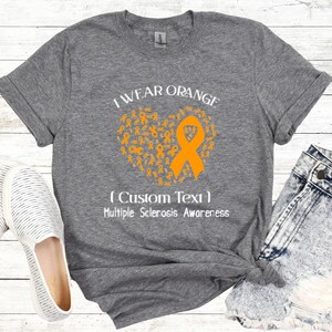 Op de afbeelding: Grijs t-shirt met de tekst "I Wear Orange" en een hartvormig ontwerp gemaakt van oranje linten. Het shirt bevat ook de tekst "(Custom Text) Multiple Sclerosis Awareness". Het shirt wordt gecombineerd met een spijkerbroek en witte schoenen.