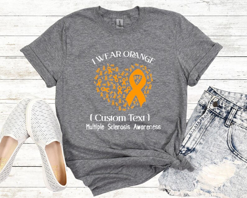 Op de afbeelding: Grijs t-shirt met de tekst "I Wear Orange" en een hartvormig ontwerp gemaakt van oranje linten. Het shirt bevat ook de tekst "(Custom Text) Multiple Sclerosis Awareness". Het shirt wordt gecombineerd met een spijkerbroek en witte schoenen.