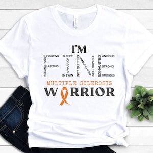 May include: White t-shirt with the text "I'M FINE" composed of words like "fighting" and "exhausted." Below, it reads "MULTIPLE SCLEROSIS WARRIOR" with an orange ribbon. The shirt is folded, with a pair of dark blue jeans visible.