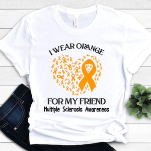 Op de afbeelding: Wit t-shirt met de tekst "I wear orange for my friend" en "Multiple Sclerosis Awareness". Het ontwerp toont een oranje lint in de vorm van een hart, samengesteld uit kleinere oranje linten.