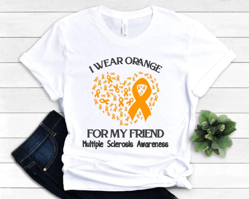 Op de afbeelding: Wit t-shirt met de tekst "I wear orange for my friend" en "Multiple Sclerosis Awareness". Het ontwerp toont een oranje lint in de vorm van een hart, samengesteld uit kleinere oranje linten.