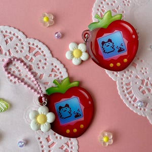 Op de afbeelding: Twee rode, aardbeivormige digitale huisdier-speelgoedjes met groene bladeren en kleine schermen met gepixeleerde karakters. Eén is bevestigd aan een roze kralenketting en een witte bloemhanger. De andere heeft een doorzichtige plastic hanger. De achtergrond bevat witte kanten placemats, kleine bloemen en vlinders.