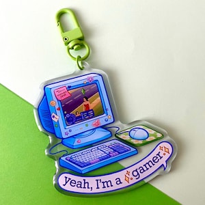 Puede incluir: Un llavero acrílico transparente con un ordenador azul con teclado, ratón y el texto "Yeah, I'm a gamer". La pantalla del ordenador muestra una escena de videojuego. El llavero tiene un cierre verde.