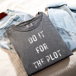Puede incluir: Una camiseta gris con el texto "DO IT FOR THE PLOT" impreso en blanco. La camiseta está doblada y colocada encima de un par de pantalones vaqueros azul claro.