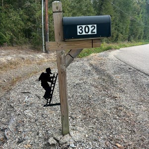 May include: A black mailbox with the number 302 in white is mounted on a wooden post. A decorative black metal silhouette of a firefighter climbing a ladder is attached to the post. The mailbox is set up near a road.