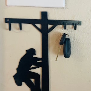 May include: Black metal key holder shaped like a lineman climbing a utility pole. The holder has multiple hooks for hanging keys.