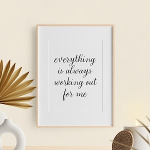 May include: A framed print with the text "everything is always working out for me" in black cursive font on a white background. The print is sitting on a wooden shelf with a vase of dried palm leaves and a woven coaster.