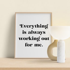 May include: A framed print with the text "Everything is always working out for me."  The print is sitting on a wooden shelf next to a white and beige table lamp.