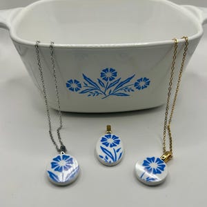 May include: Three necklaces with round, white pendants featuring a blue floral design. The necklaces have silver and gold chains. A white casserole dish with a blue floral pattern is in the background.