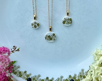 Pode incluir: Três colares de corrente dourada com pingentes circulares brancos com um padrão floral verde. Os pingentes são exibidos num prato branco com uma borda floral verde a condizer, entre cachos de flores cor-de-rosa e brancas.