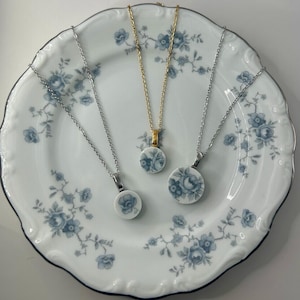 May include: Three necklaces with round, porcelain pendants featuring blue floral designs. The pendants are on silver and gold chains. The necklaces are displayed on a white plate with a blue floral pattern and a silver rim.