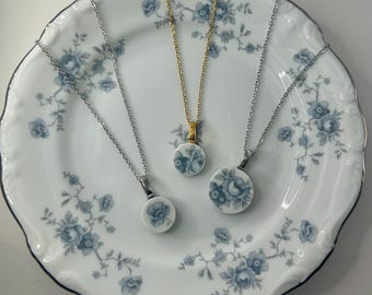 Pode incluir: Três colares com pingentes redondos de porcelana com desenhos florais azuis. Os pingentes estão em correntes de prata e ouro. Os colares são exibidos em um prato branco com um padrão floral azul e uma borda prateada.