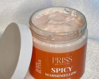 May include: A clear plastic jar filled with pale peach-colored body butter, featuring an orange label with white text that reads PRISS BODY COLLECTION SPICY MARSHMELLOW VEGAN ORGANIC. The jar has a white lid resting beside it on the left side.