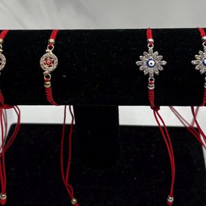 May include: Six red string bracelets with decorative silver charms are displayed on a black velvet stand. The charms include stars, circles, and a blue eye design. The bracelets are on a white background.