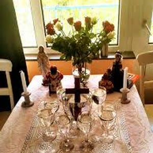 May include: A white tablecloth with a lace pattern covers a table with a wooden cross, six clear glass goblets, two white candles, and three religious figurines. The figurines are a woman in a white dress, a man in a red and gold robe, and a woman in a red and black dress. A bouquet of red and white roses is in a clear glass vase.